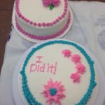 2 cakes. one with white frosting and pink piping and pink flowers, the other with pink flowers, blue piping and the words "I did it!" on the top